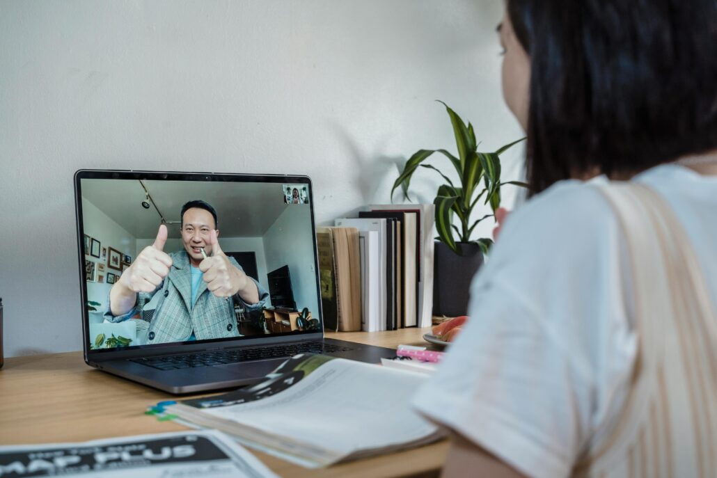 A coach and client communicate through a video call on a laptop. The coach on the screen is smiling and giving two thumbs up, while the client sits at a desk with notebooks, a laptop, and books nearby, symbolizing an online coaching session and positive connection.