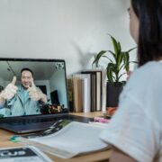 A coach and client communicate through a video call on a laptop. The coach on the screen is smiling and giving two thumbs up, while the client sits at a desk with notebooks, a laptop, and books nearby, symbolizing an online coaching session and positive connection.