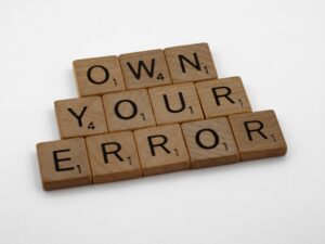 Scrabble tiles spelling “Own Your Error” on a wooden surface, symbolizing personal responsibility, growth, and the power of accountability in coaching.