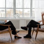 A life coach and client sit facing each other in a bright, minimalist office with wooden floors and large windows. The coach, holding a notebook, listens attentively as the client gestures while speaking, illustrating a focused one-on-one coaching session.