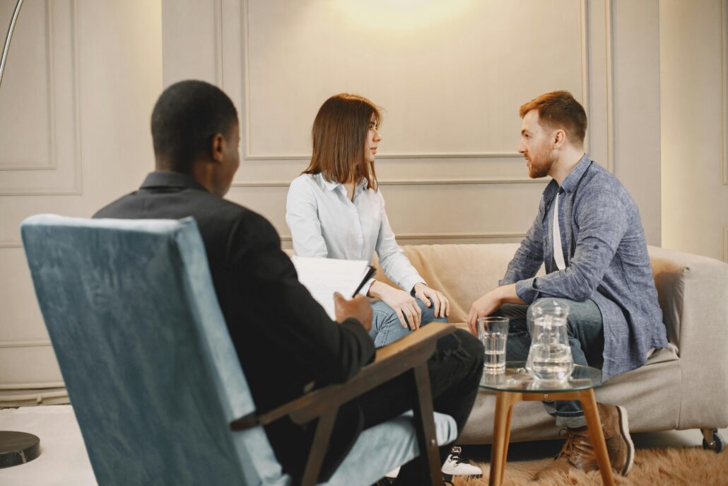 A couple sits on a beige couch during a relationship coaching session, attentively speaking with a coach seated across from them taking notes in a modern, warmly lit office.