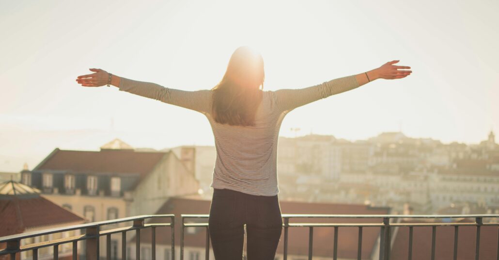 A woman stands on a balcony with her arms outstretched, facing the morning sun over a city skyline. The warm sunlight creates a glowing, hopeful atmosphere, symbolizing confidence, freedom, and self-empowerment — aligning with the theme of overcoming imposter syndrome and embracing authenticity.