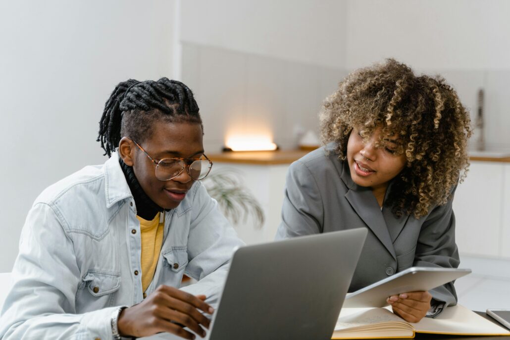 Two people sitting together at a desk having a focused coaching conversation. One person is using a laptop while the other holds a tablet and offers guidance, both smiling and engaged in a collaborative discussion.