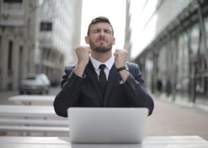 A determined man in a business suit sitting at a laptop outdoors, clenching his fists in a moment of emotional resilience or triumph, symbolizing overcoming setbacks through inner strength.