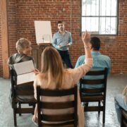 Group coaching session with a diverse group of adults, one participant raising her hand while the coach leads a discussion in a brick-walled room.