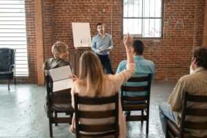 Group coaching session with a diverse group of adults, one participant raising her hand while the coach leads a discussion in a brick-walled room.