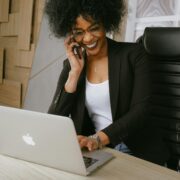 Smiling woman sitting in front of a laptop, talking on her phone, celebrating progress—symbolizing how to help a stuck client move forward.